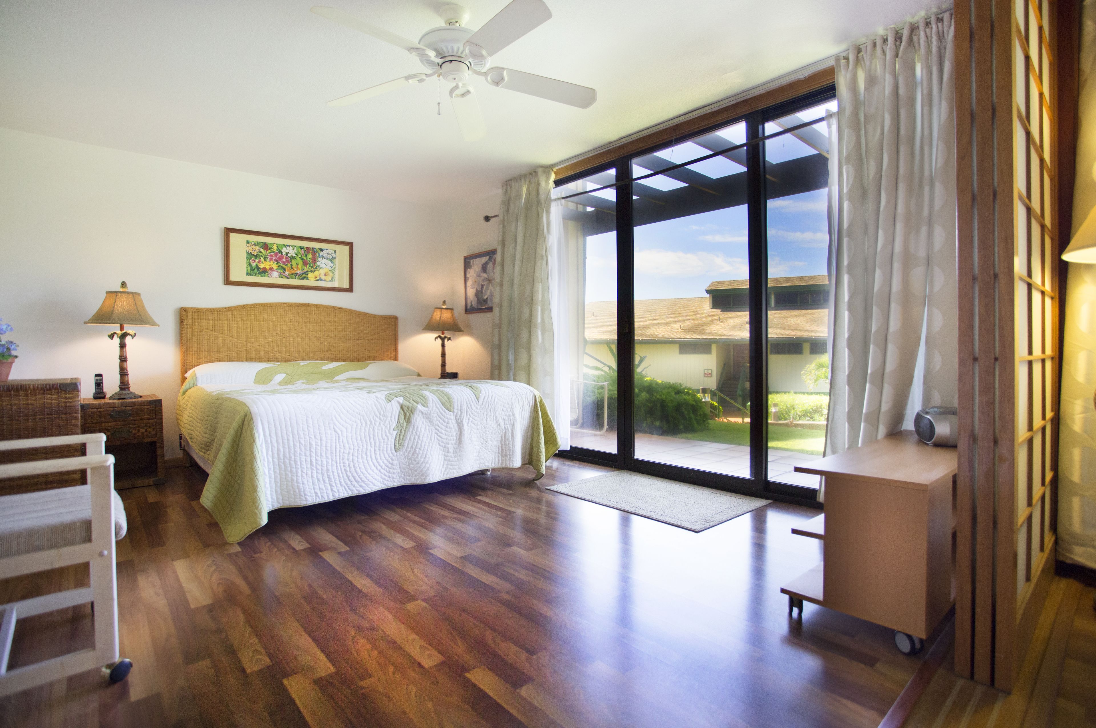 A bedroom with a bed, two bedside tables, a ceiling fan, a window with curtains, and a view of the outside.
