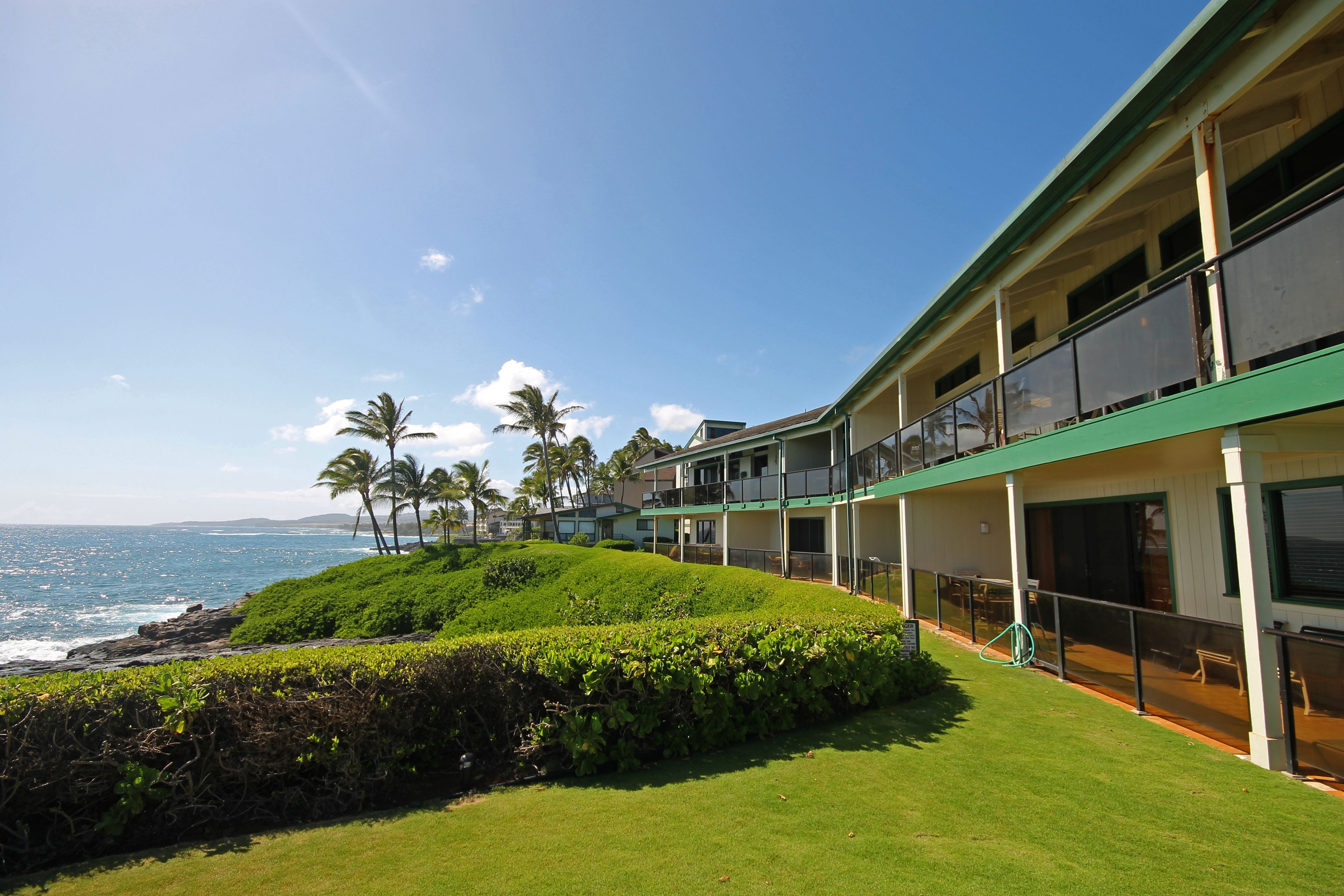 A hotel with a balcony overlooking the ocean, a grassy area, and palm trees.