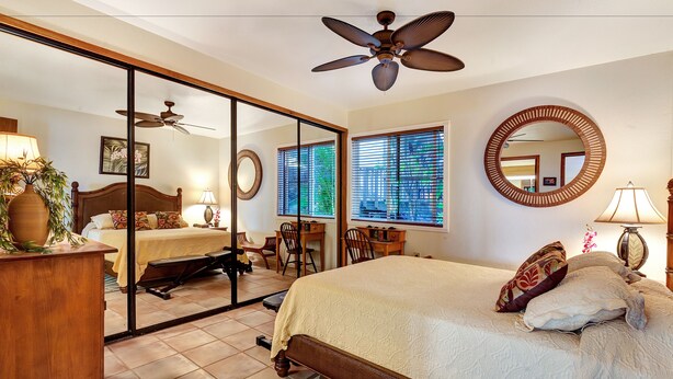 A bedroom with a bed, a desk, a chair, a ceiling fan, and a large mirror. - Castle at Makahuena at Poipu (Koloa)
