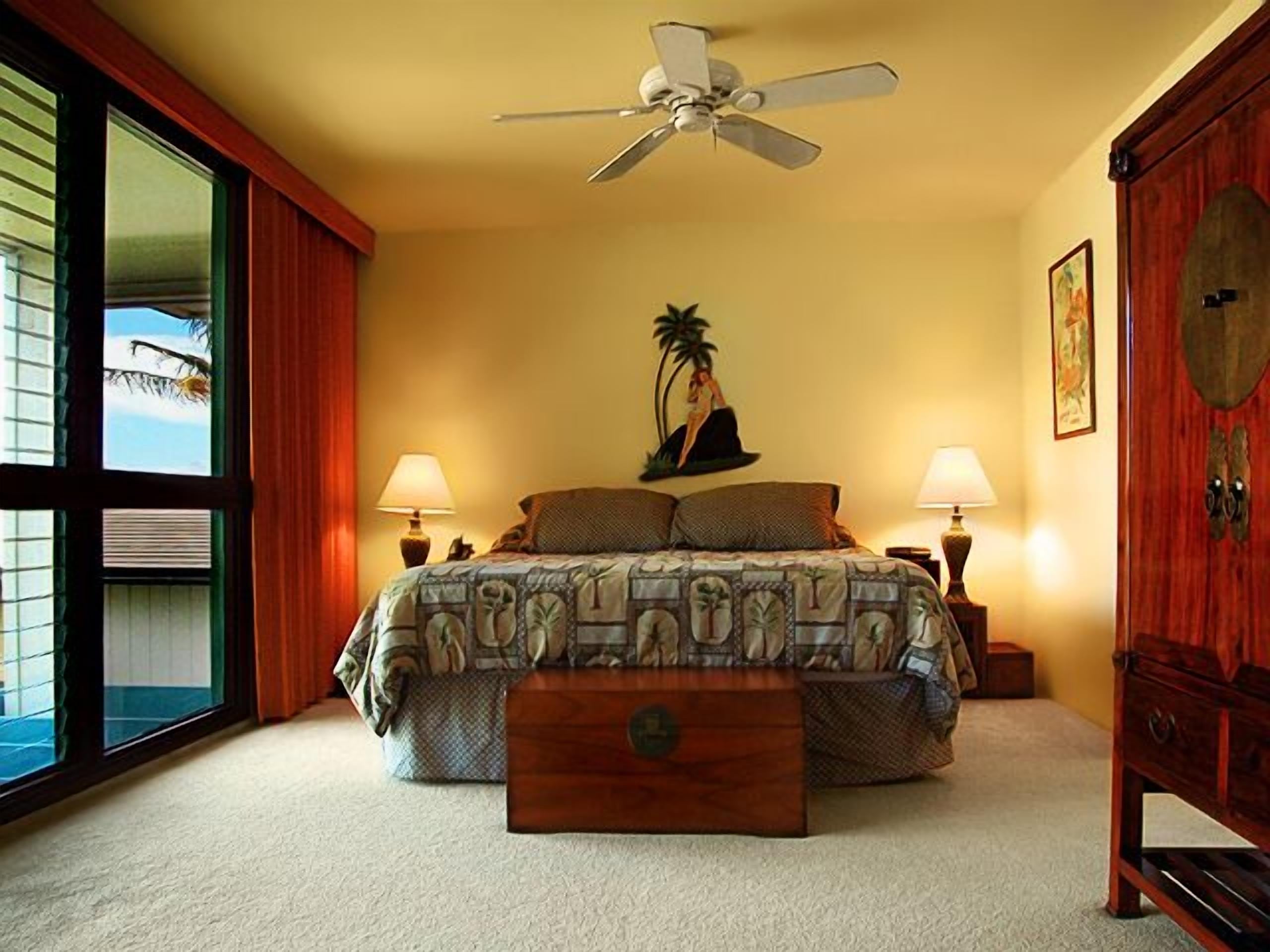 Une chambre à coucher comprenant un lit, deux lampes de chevet, un ventilateur de plafond, une table de chevet en bois, une commode en bois et une grande fenêtre avec des stores.