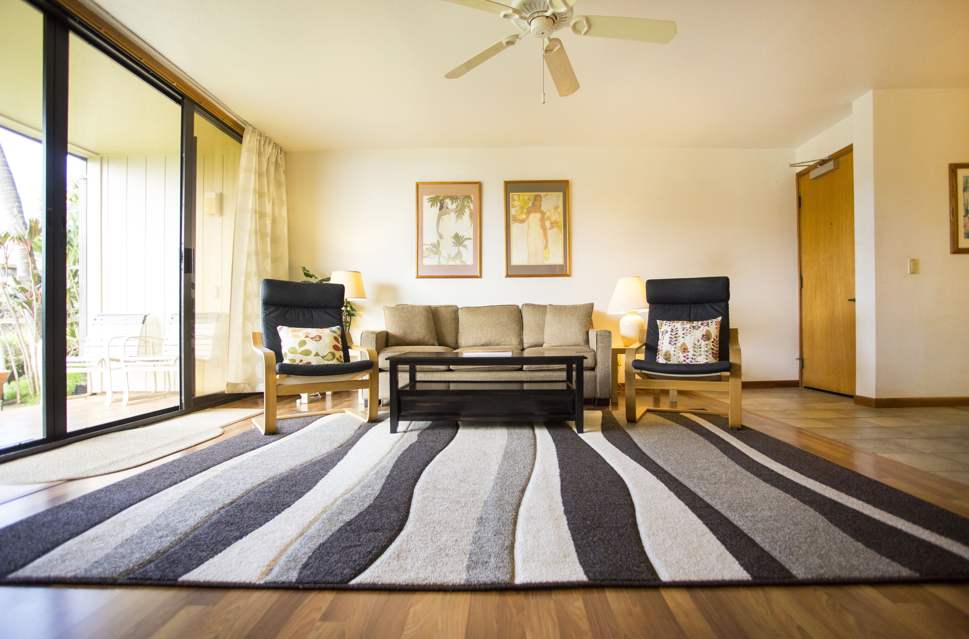 A living room with a sofa, two armchairs, a coffee table, and a ceiling fan. There are two framed pictures on the wall and a sliding glass door leading to an outdoor area.