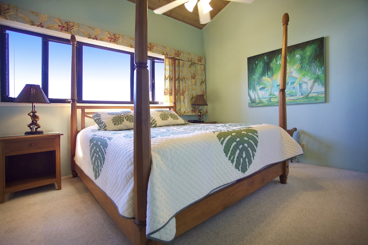 A four-poster bed with a leaf-patterned bedspread, a nightstand with a lamp, and a painting of palm trees on the wall.