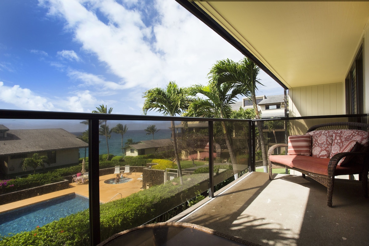 A balcony with a wicker sofa, a pool, and a view of the ocean and palm trees.