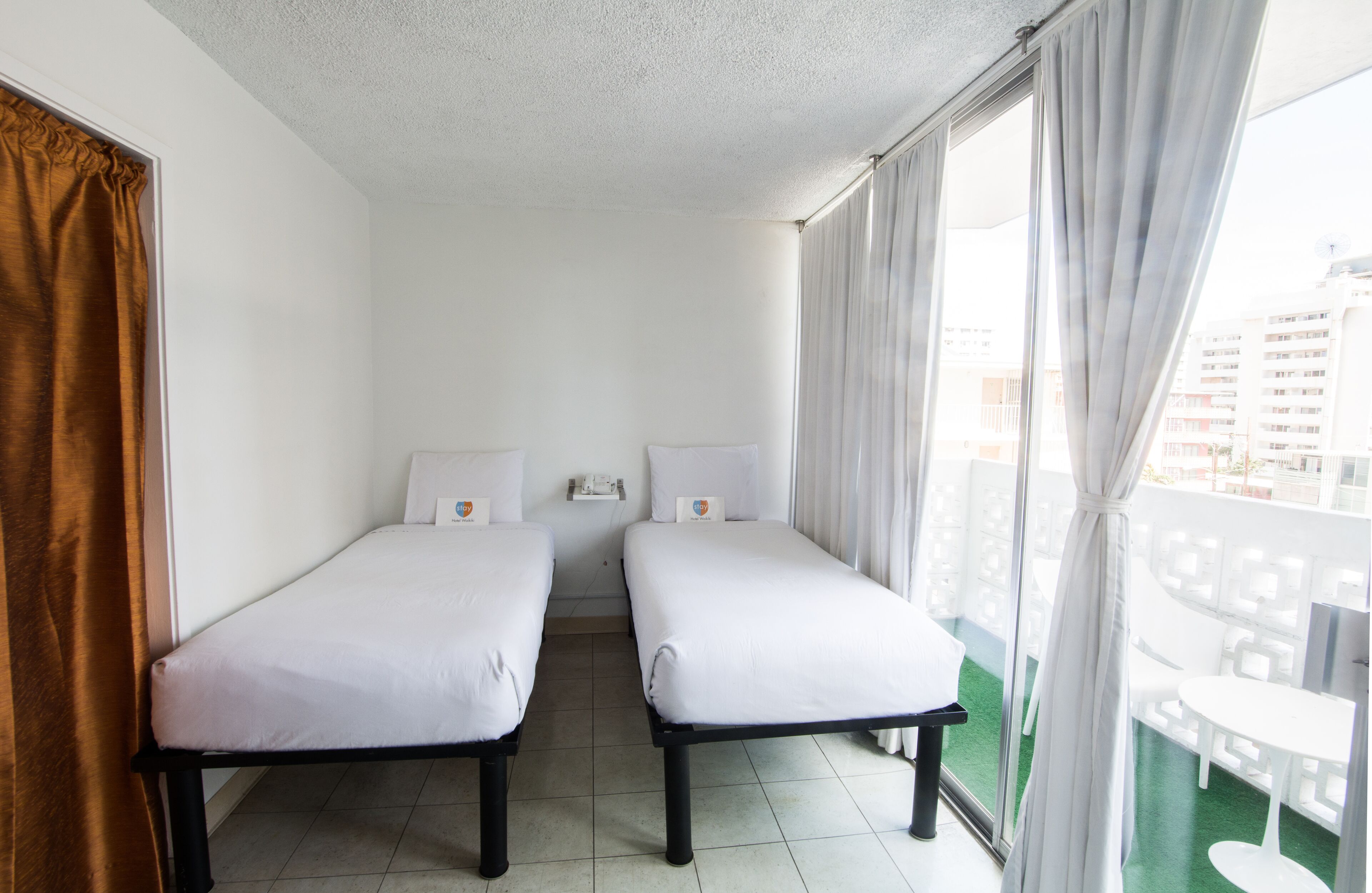 A hotel room with two single beds, a window with curtains, a view of the city, and a balcony with white furniture.