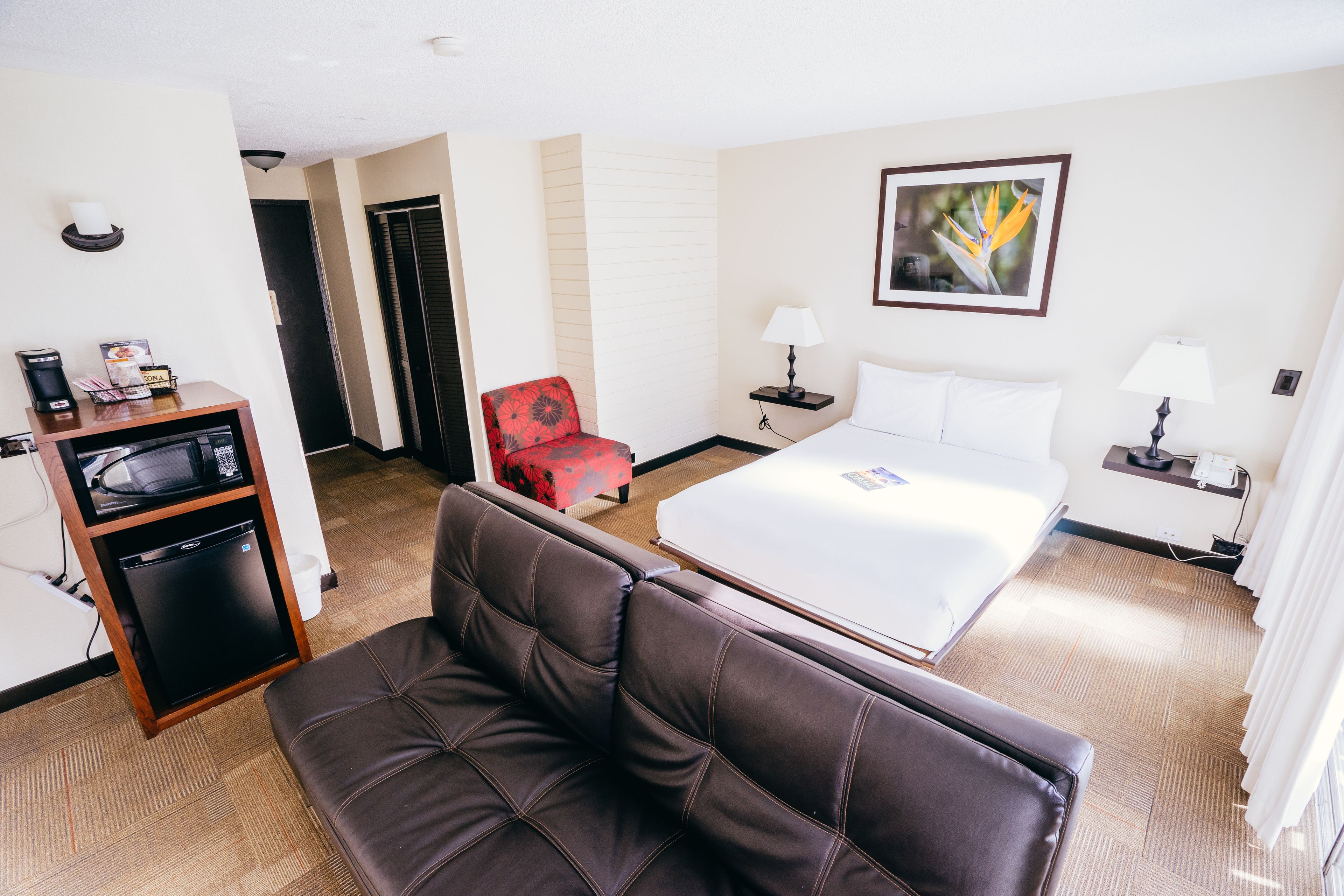 Een hotelkamer met een bed, een bruine leren bank, een rode fauteuil, een klein bureau met een lamp en een foto aan de muur.