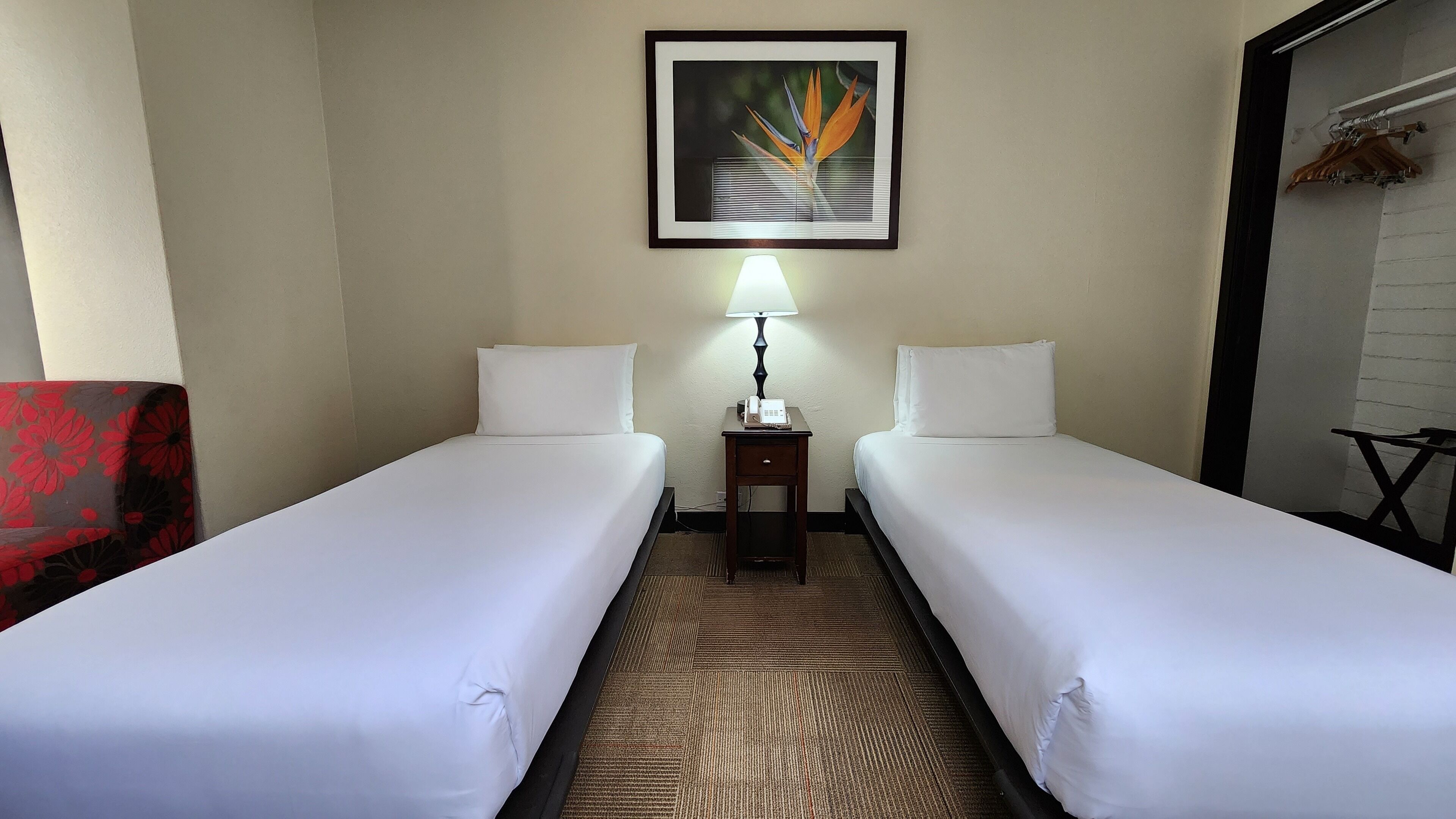 A hotel room with two single beds, a bedside table with a lamp, a framed picture on the wall, and a red chair.