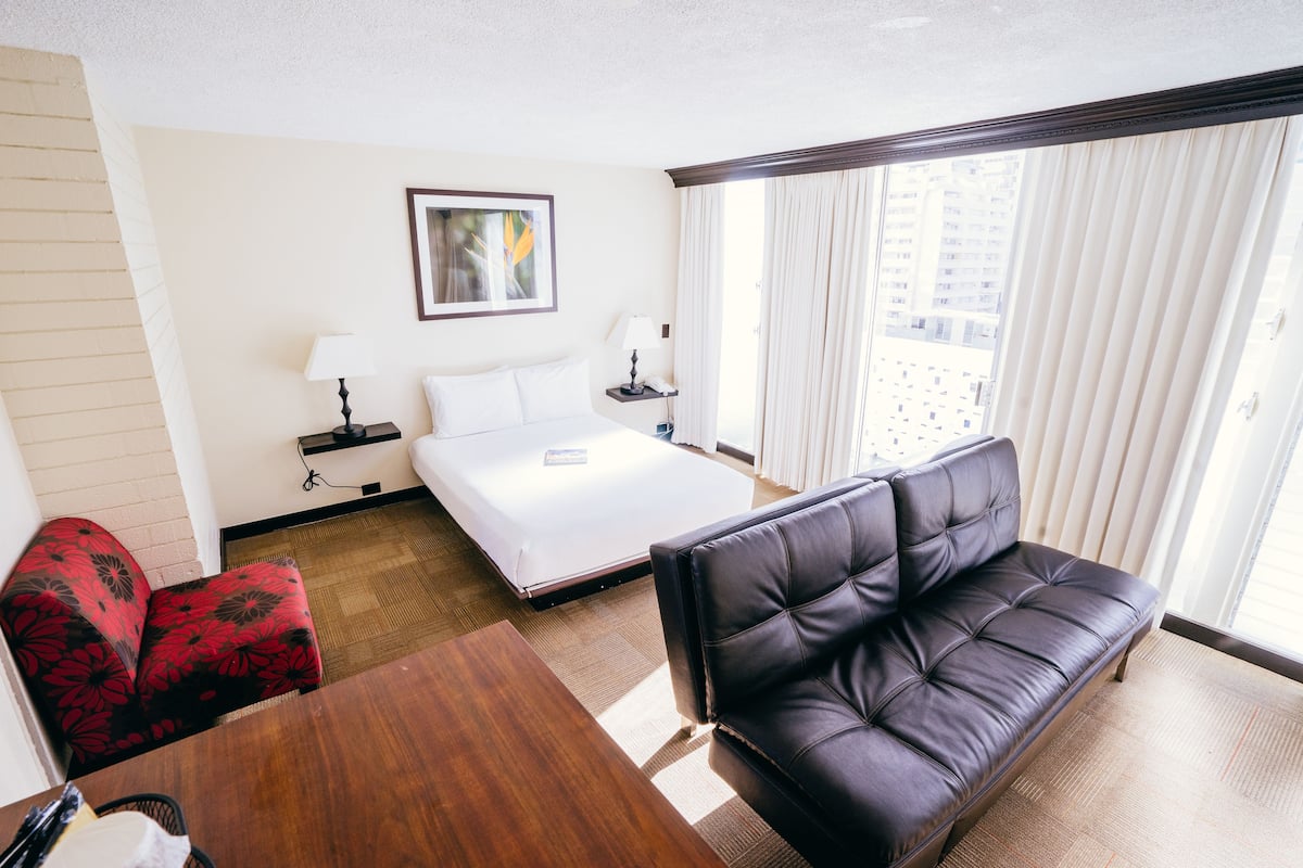 A hotel room with a bed, a leather sofa, a wooden table, a red patterned chair, a nightstand with a lamp, and a window with curtains.
