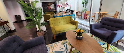 A hotel lobby with a seating area featuring a sofa, armchairs, and a coffee table. There are potted plants and a large window providing a view of the outside.
