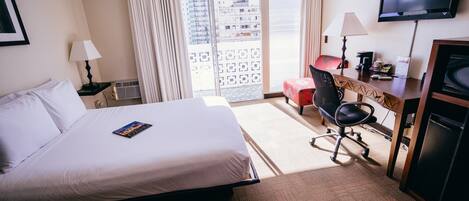 A hotel room with a large bed, a desk with a computer, a TV, and a view of the city through a balcony.