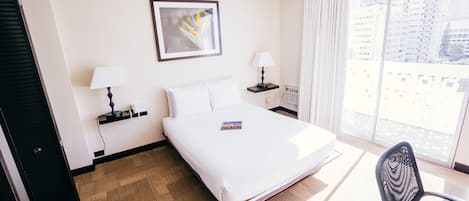 Habitación de hotel con cama, dos mesitas de noche con lámparas, una silla, una ventana con vistas a edificios y una puerta.