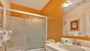 A bathroom with a glass shower enclosure, a white toilet, a sink with toiletries, and a framed picture on the wall.