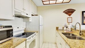 A kitchen with white cabinets, a microwave, stove, refrigerator, granite countertop, sink, and a decorative fan on the wall. - Castle at Kona Reef (Kailua-Kona)