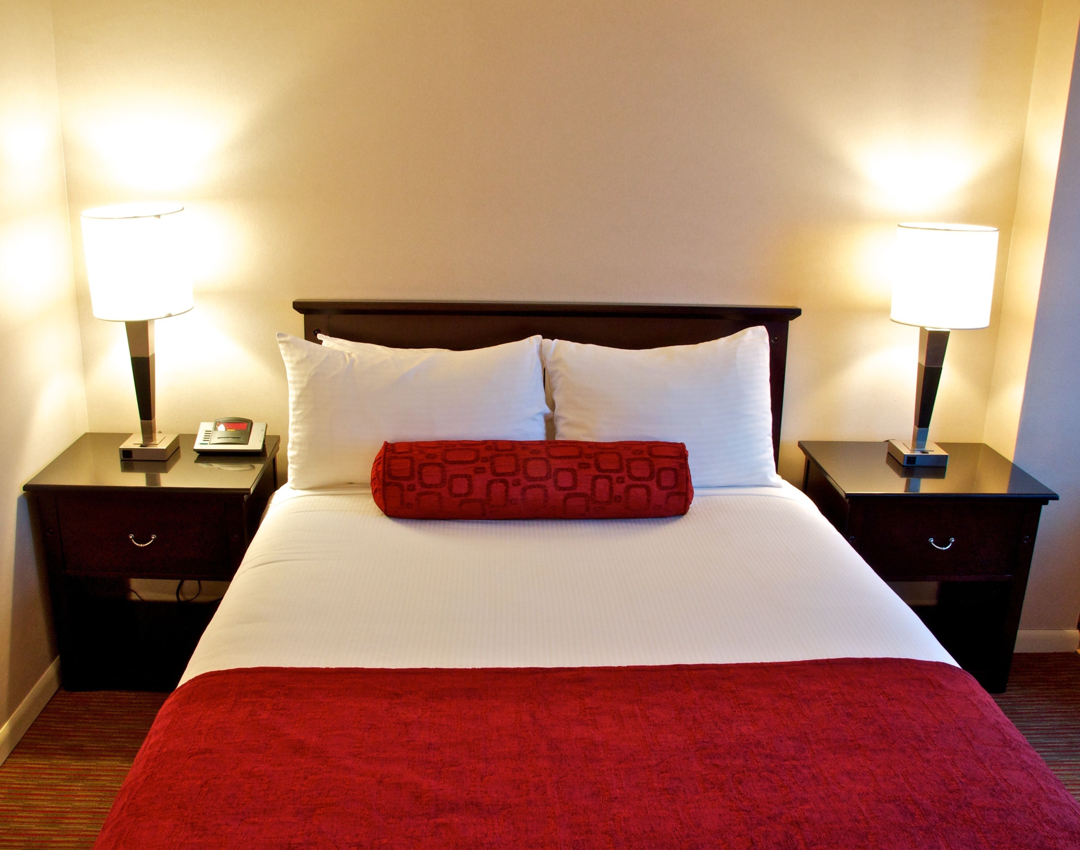 A hotel room with a bed, two bedside tables, two lamps, and a red throw pillow.