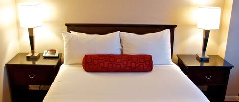 Habitación de hotel con cama, dos mesitas de noche, dos lámparas y un cojín rojo.