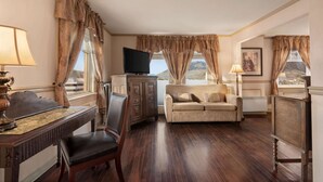 A room with a wooden floor, a television, a sofa, a desk with a lamp, and curtains. - The Plaza Hotel Downtown, Trademark Collection by Wyndham (Kamloops)