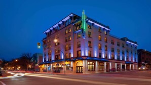 A multi-story hotel with a prominent green vertical light feature, multiple ground-level entrances, and a well-lit facade. - The Plaza Hotel Downtown, Trademark Collection by Wyndham (Kamloops)