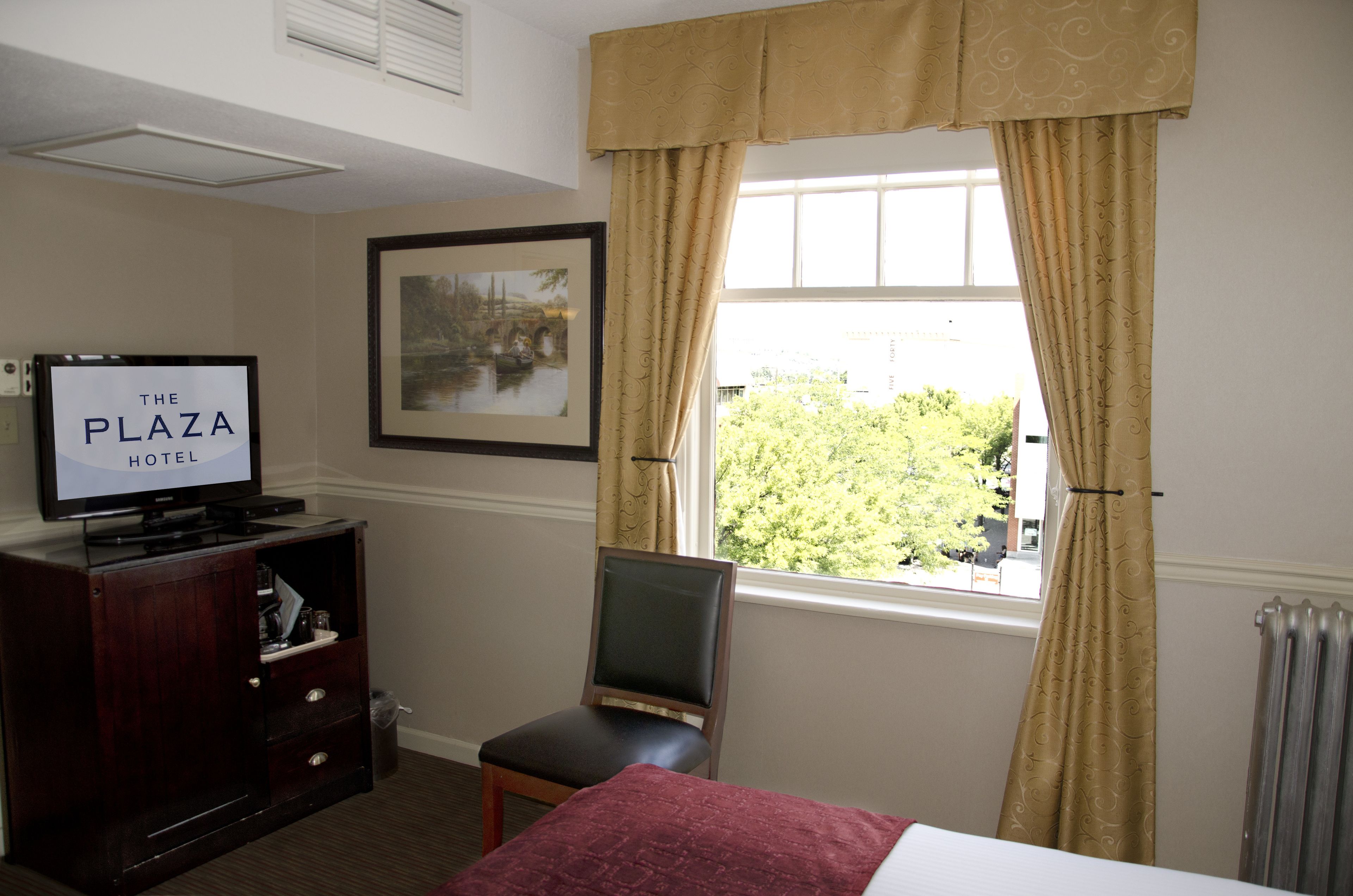 A hotel room with a bed, a television on a wooden cabinet, a chair, a window with curtains, and a framed picture on the wall.