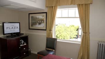 A hotel room with a bed, a television on a wooden cabinet, a chair, a window with curtains, and a framed picture on the wall.