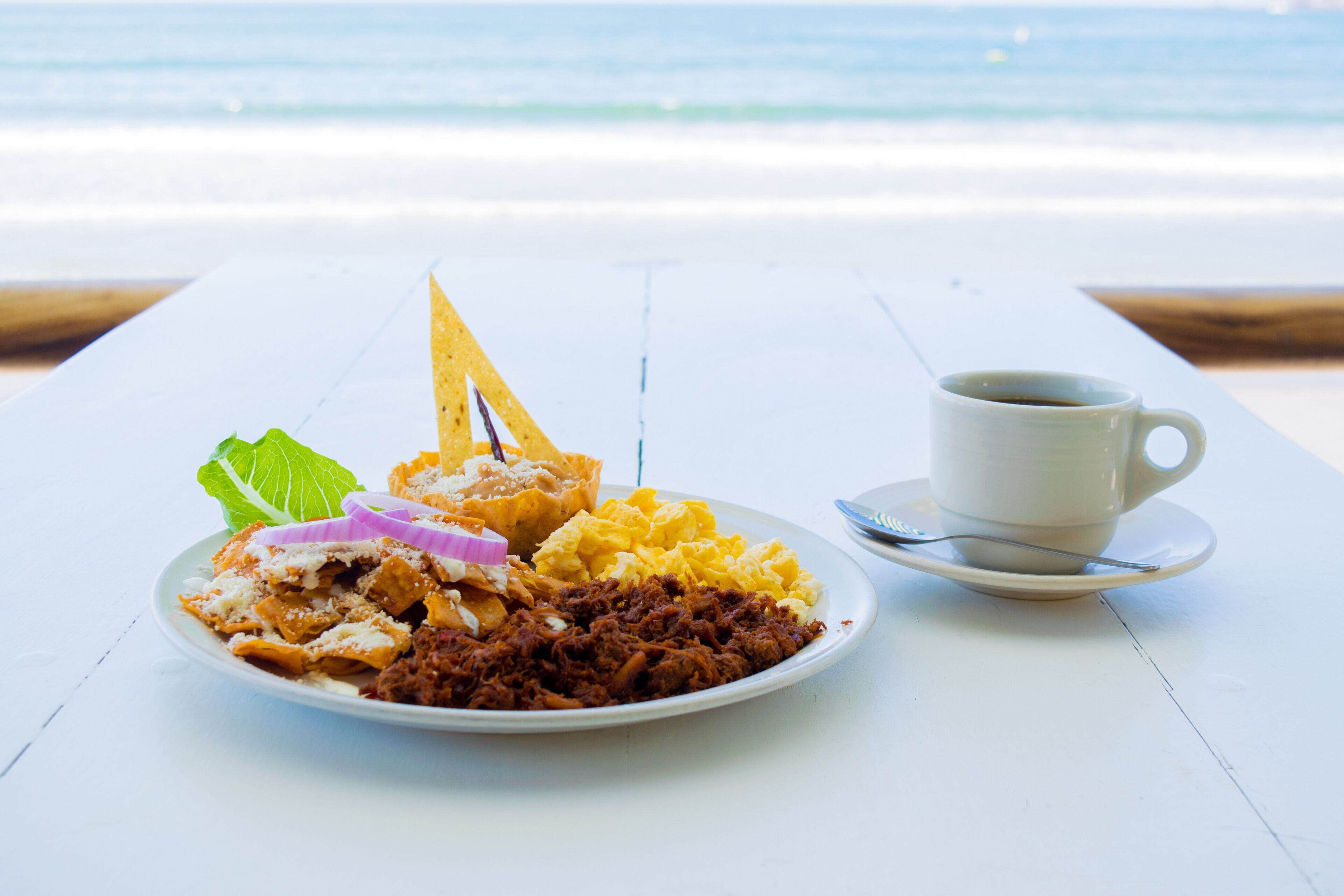 Une assiette de nourriture et une tasse de café sur une table blanche, près de la plage.