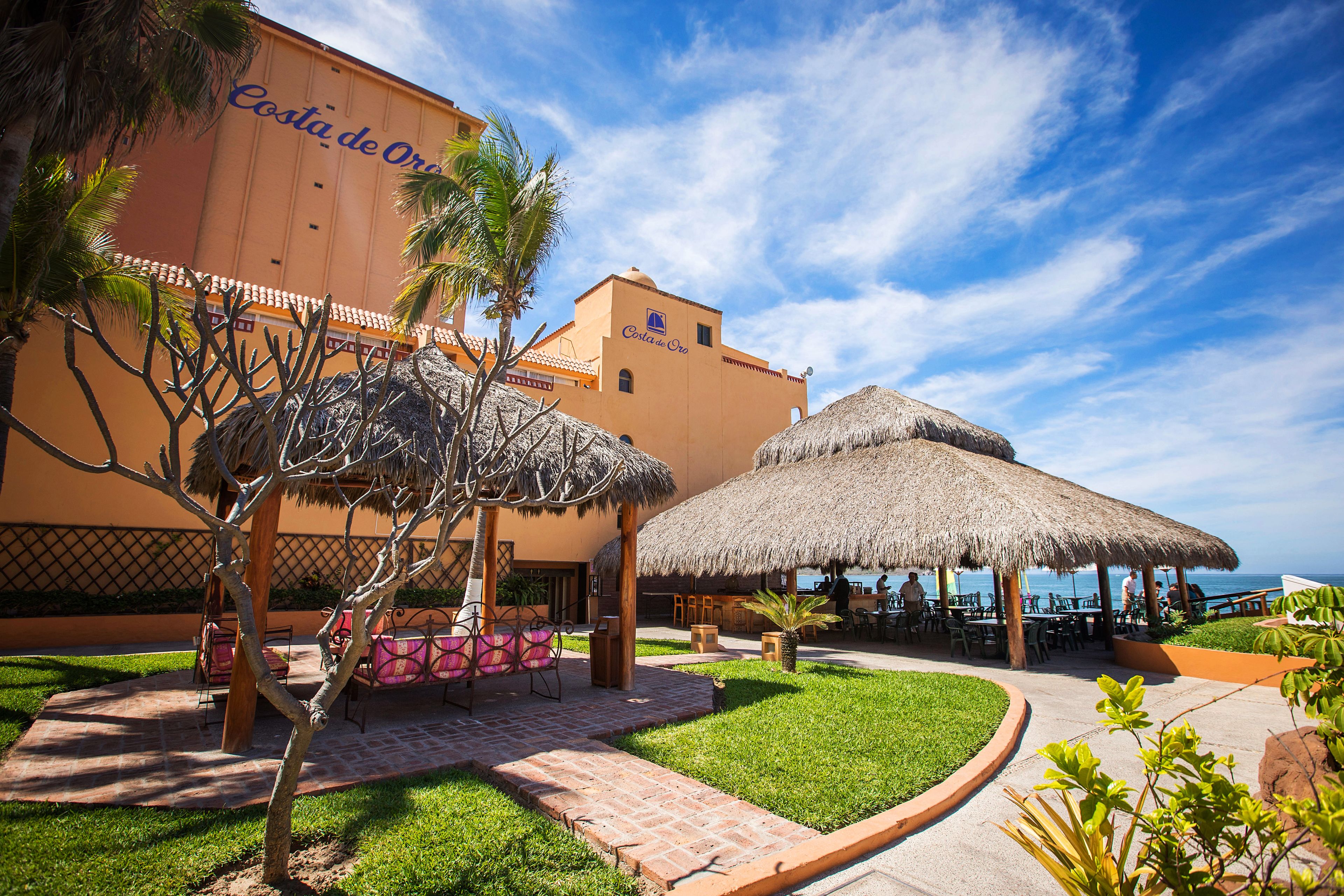A resort with a thatched-roof outdoor seating area, a pool, and a building with "Costa de Oro" signage.