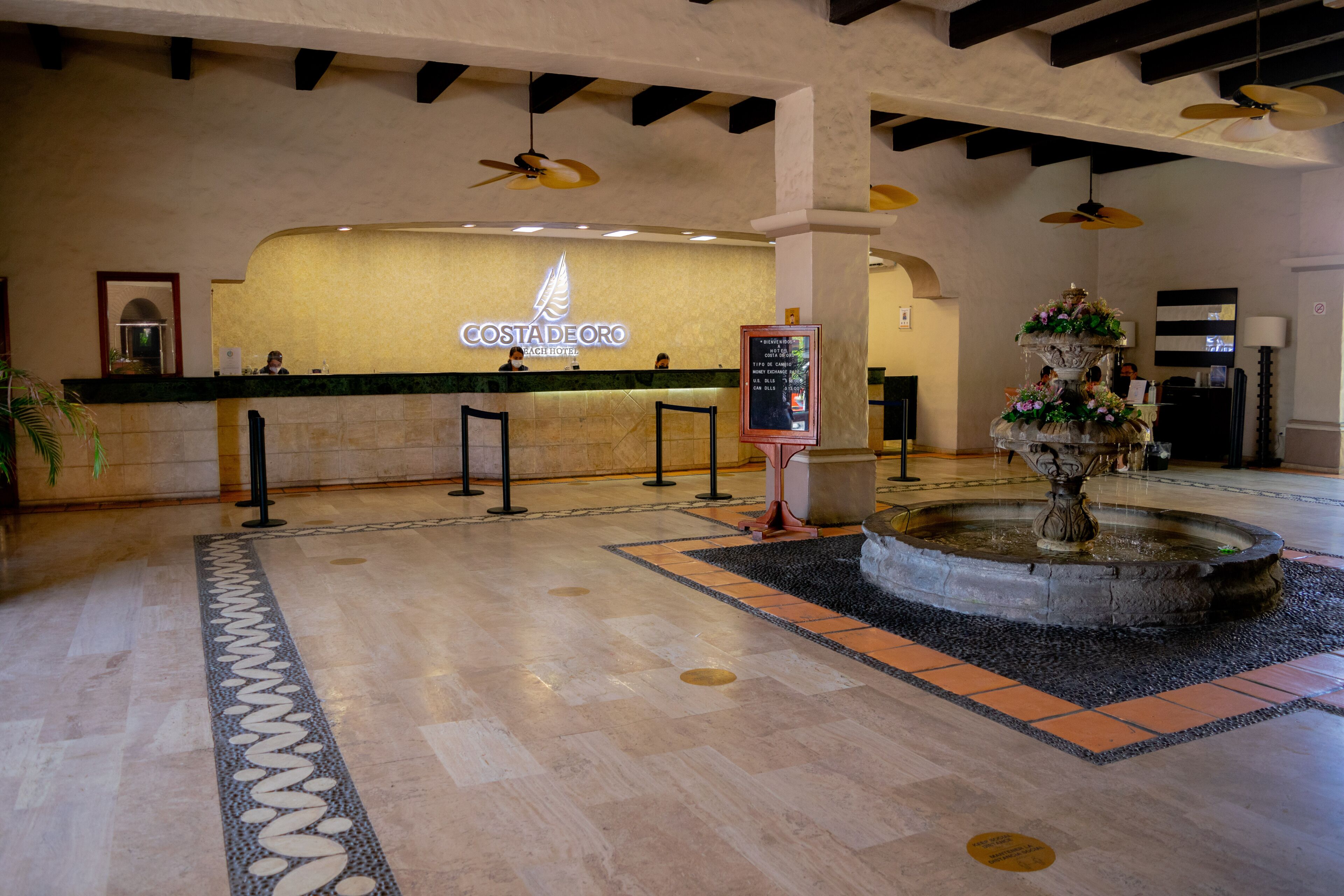 A reception area with a Costa de Oro sign, a reception desk, a fountain, and a tiled floor with a decorative border.