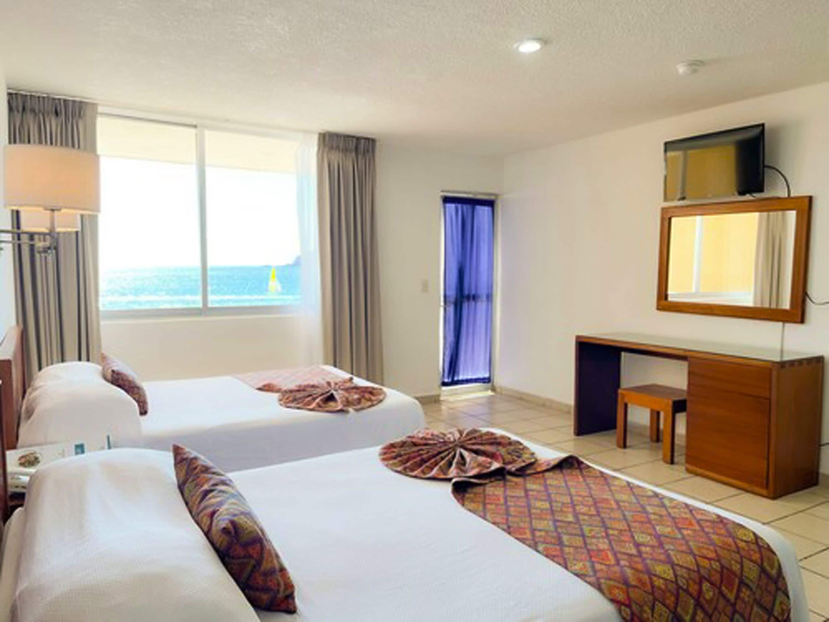 Habitación de hotel con dos camas, un escritorio, una silla, un televisor y ventana con vista al mar.