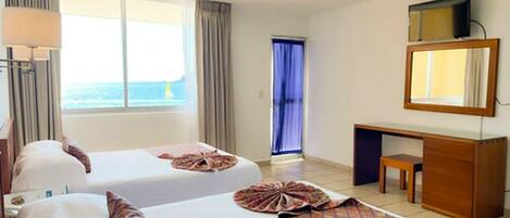 Habitación de hotel con dos camas, un escritorio, una silla, un televisor y ventana con vista al mar.