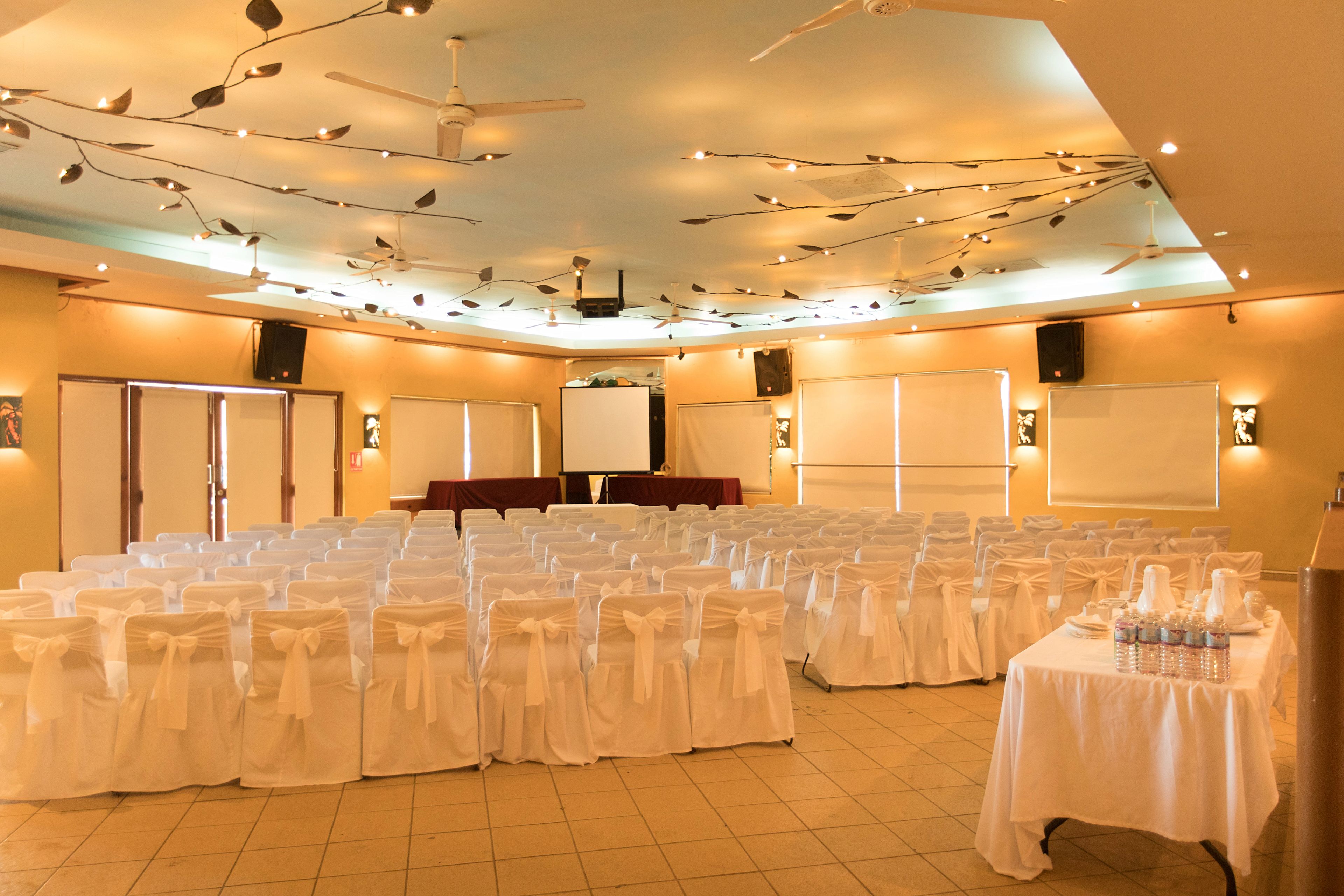 A conference room with rows of chairs covered in white, a table with water bottles, and a ceiling fan.