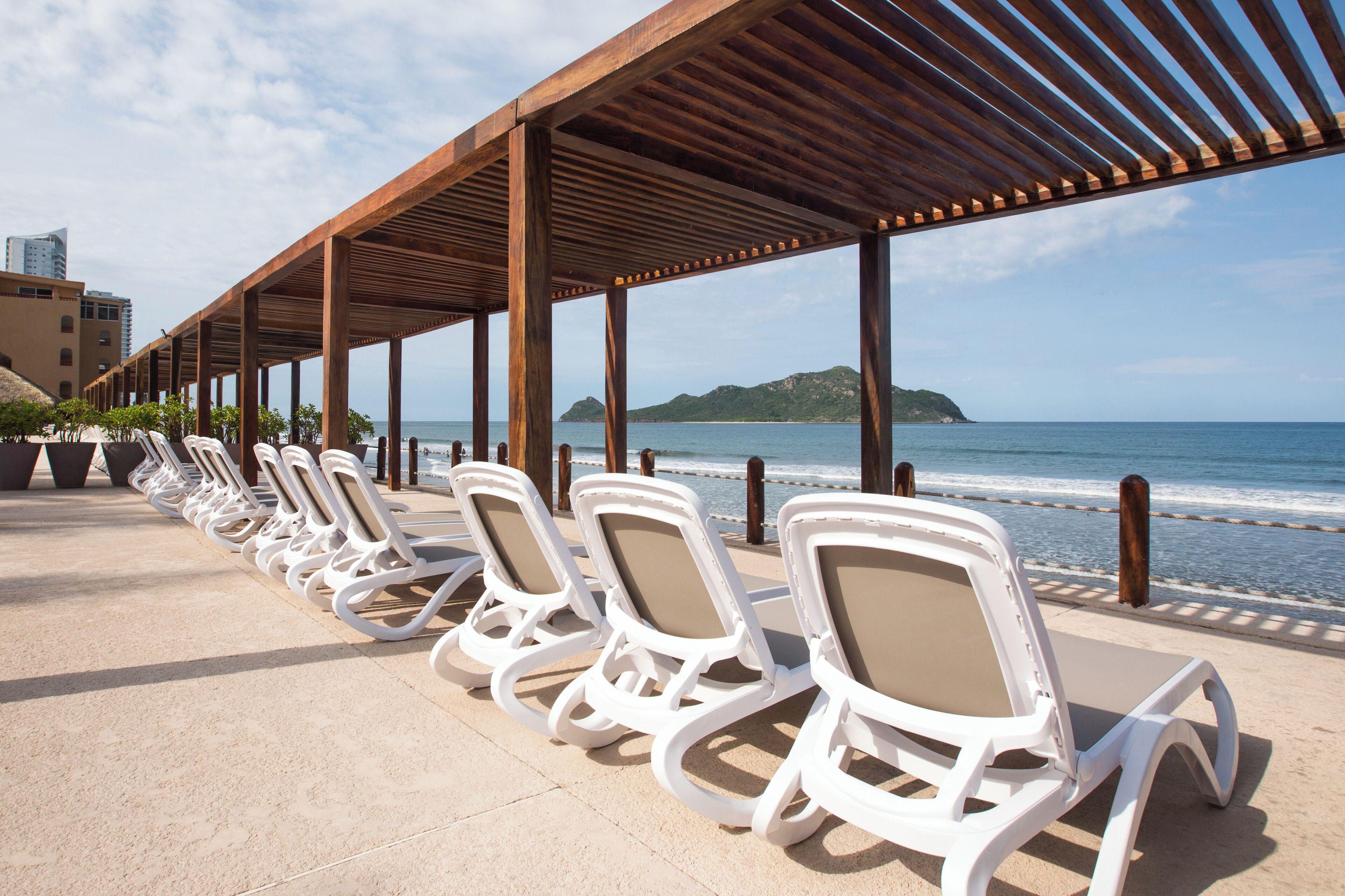 Un salón junto a la playa con hileras de sillas blancas bajo una pérgola de madera, con vistas al océano.