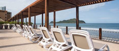 Un salón junto a la playa con hileras de sillas blancas bajo una pérgola de madera, con vistas al océano.