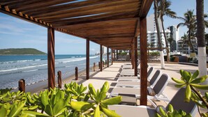 Un salon de plage doté d’une pergola en bois, de chaises longues blanches et offrant une vue sur l’océan et les palmiers.