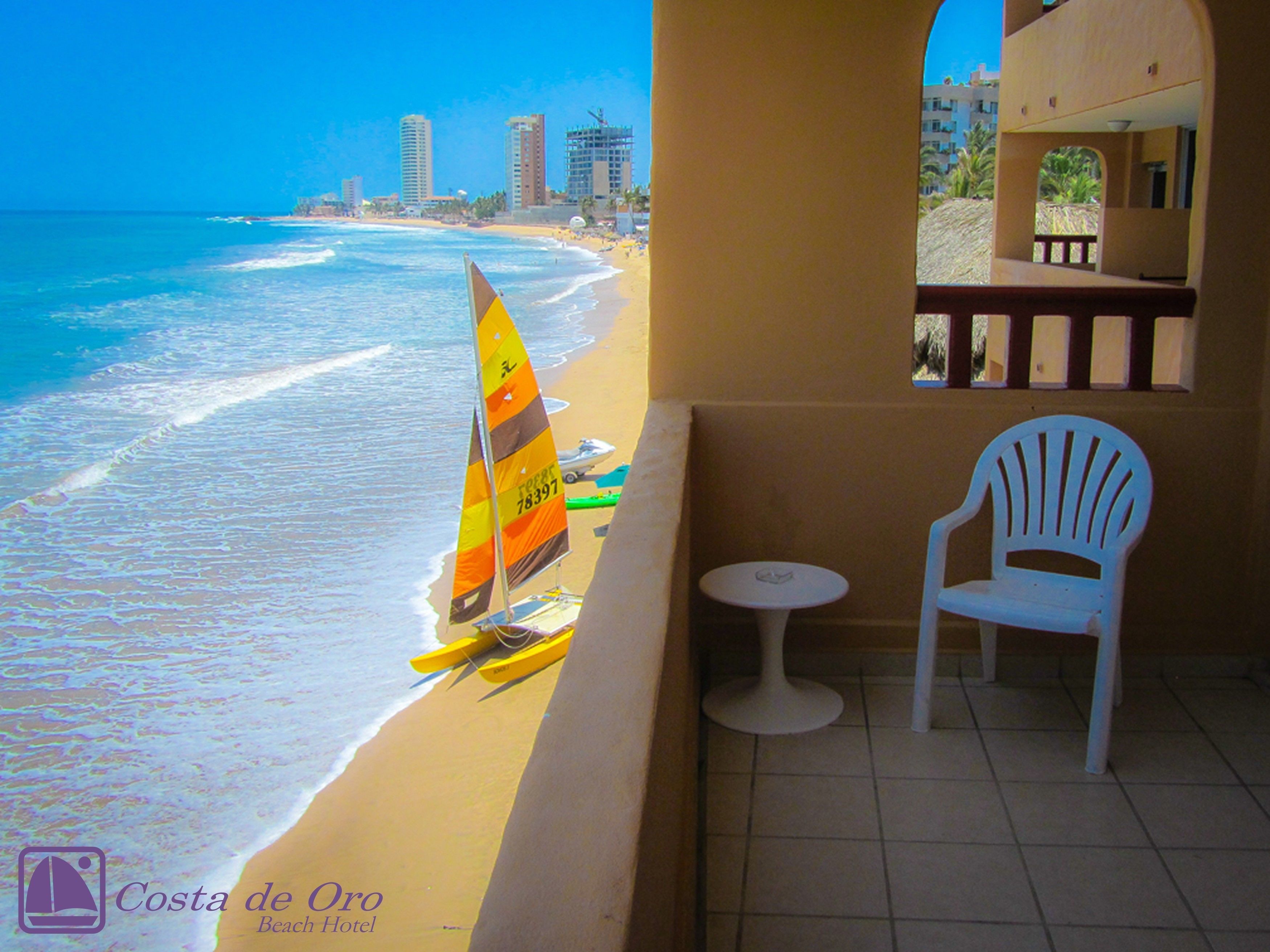 A beach view from a balcony with a chair and a small table, a sailboat on the sand, and a cityscape in the background.