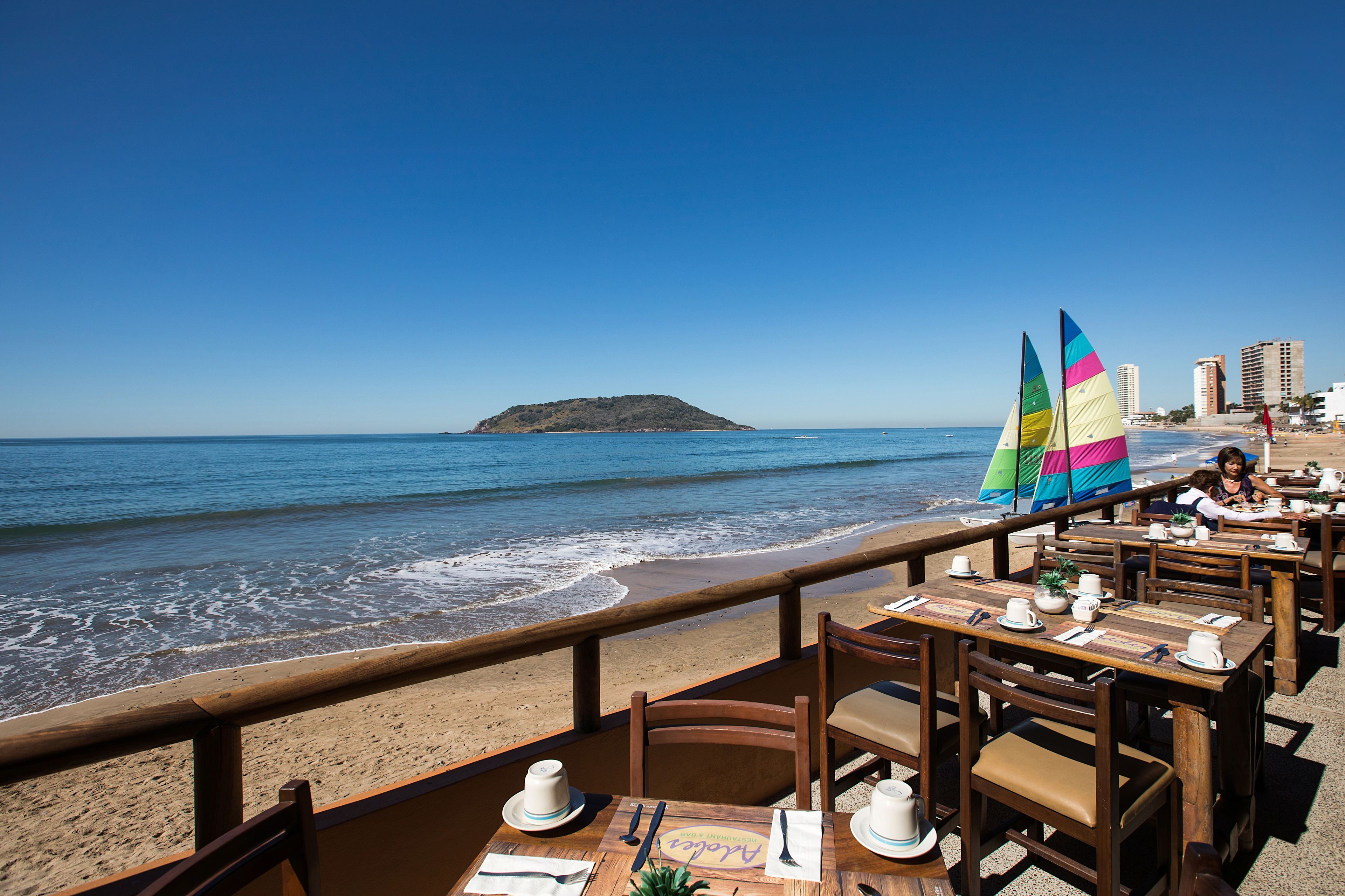 Un restaurante frente al mar con mesas y sillas de madera, vista al océano y una isla distante.