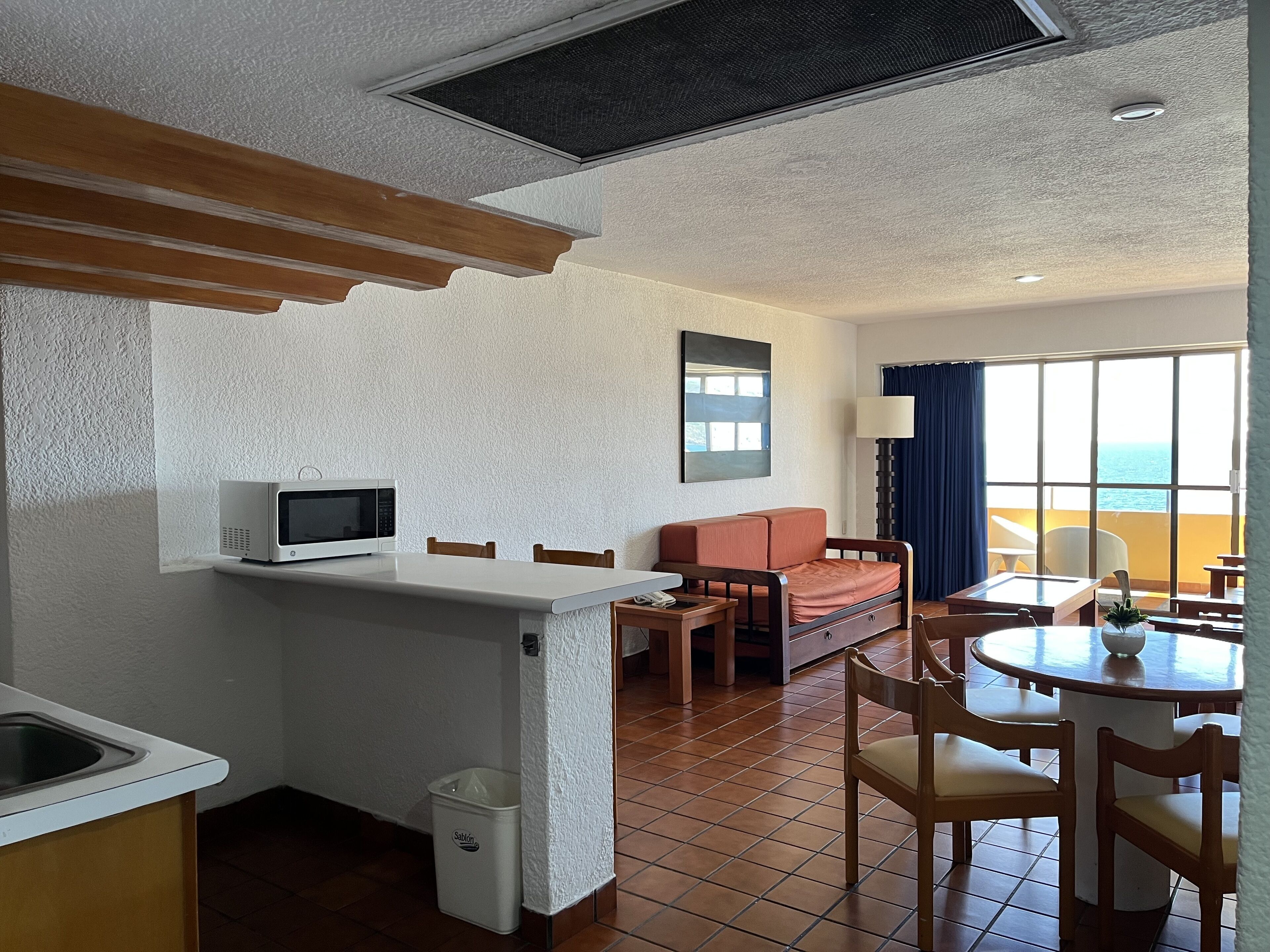Ett kompakt hotellrum med ett litet kök, en soffa, ett matbord och utsikt över havet.