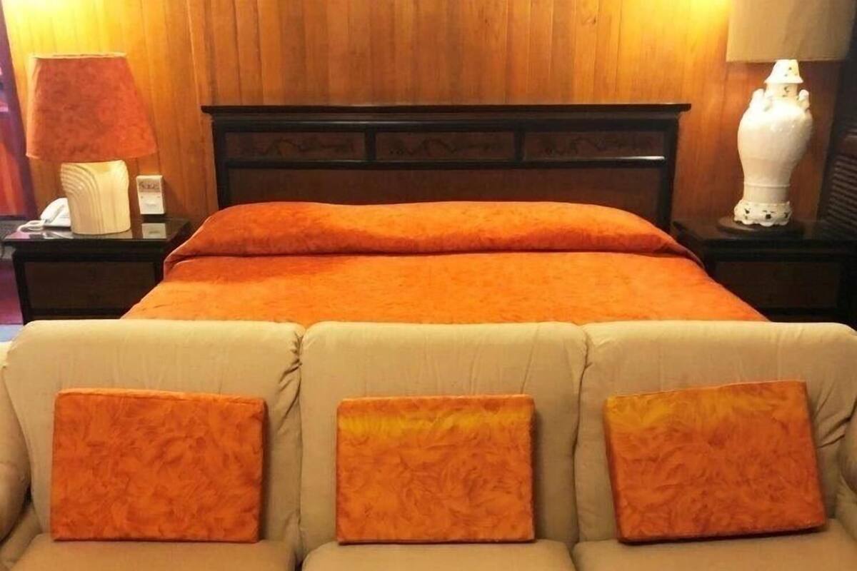 Una cama con manta naranja, cabecera de madera y sofá beige con cojines naranjas.