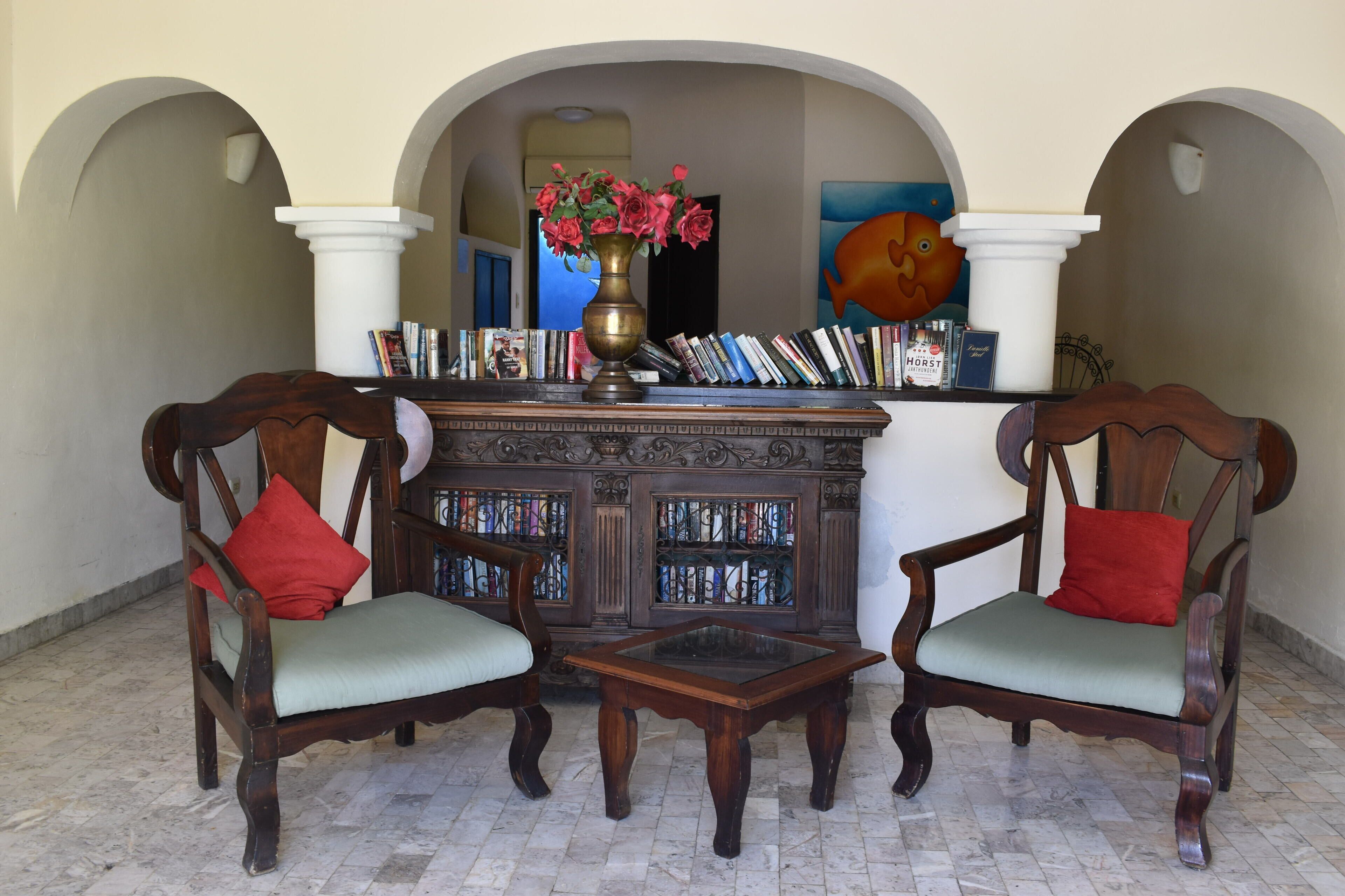 Eine Lobby mit zwei Holzsesseln, einem Couchtisch, einem Bücherregal und einer Vase mit Blumen.