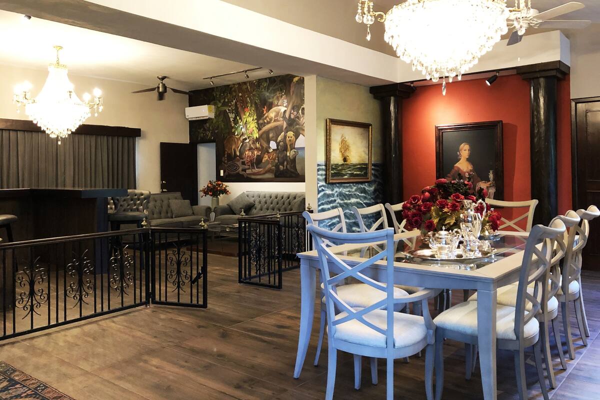 Un comedor con una mesa dispuesta para seis personas, rodeada de sillas azules, una araña y un cuadro en la pared.