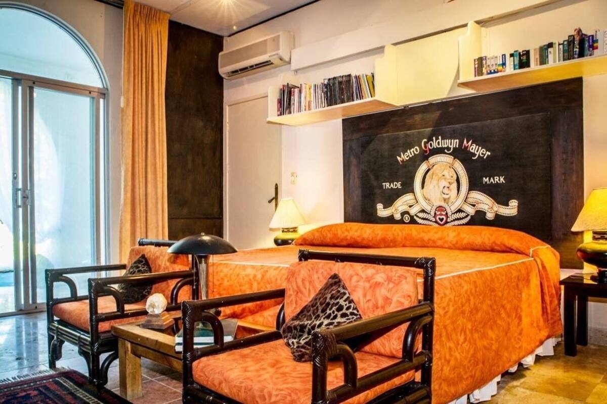 Una habitación de hotel con una cama, una silla, una mesa, una lámpara, una estantería y un ventanal amplio.