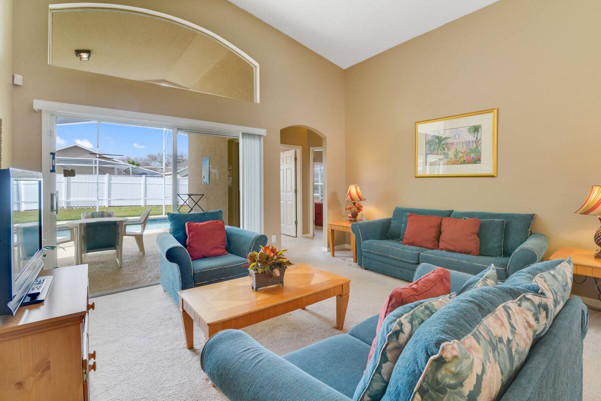Ein geräumiges Wohnzimmer mit zwei blauen Sofas, einem Holztisch und einem Fernseher. Eine Tür führt zu einem Balkon mit Blick auf Häuser.