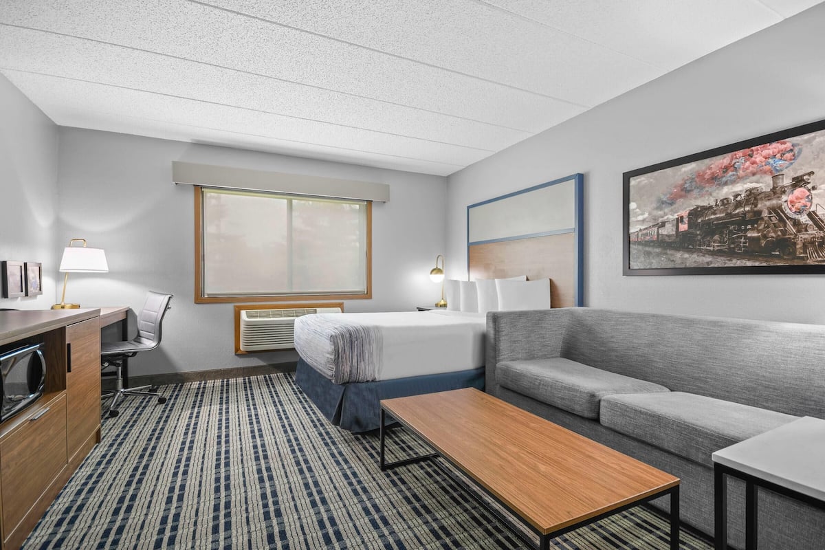 Una habitación de hotel con una cama, un sofá, un escritorio con una lámpara, un ventanal grande y un cuadro en la pared.