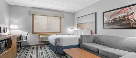 A hotel room with a bed, a sofa, a desk with a lamp, a large window, and a painting on the wall.