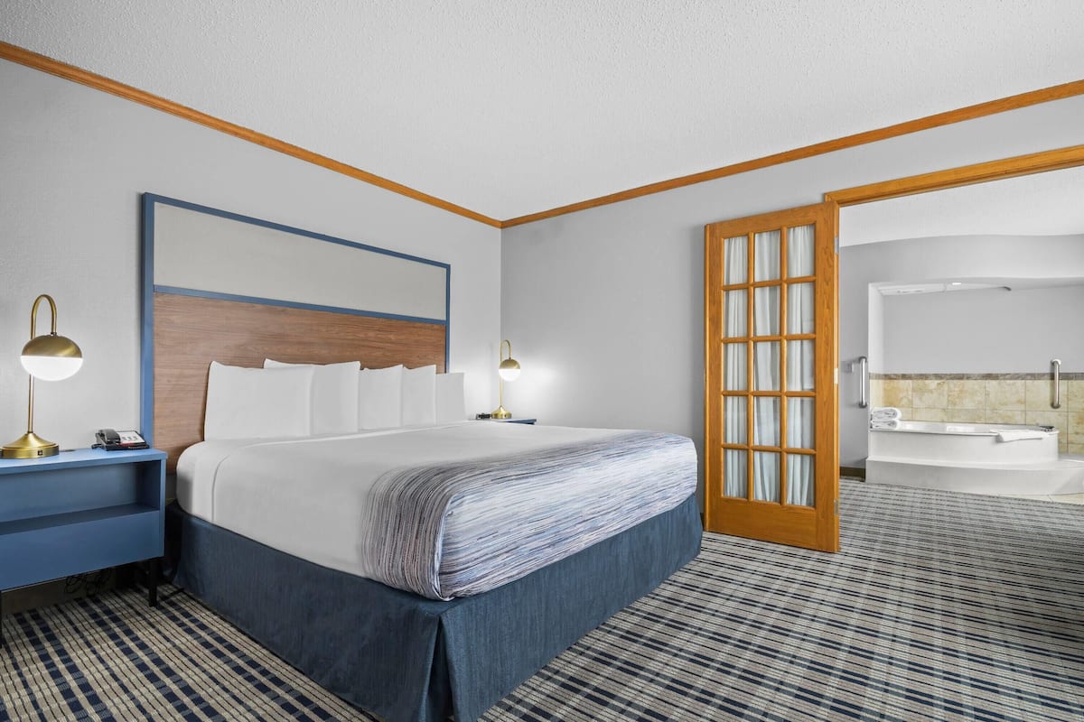 Habitación de hotel con una cama grande, lámparas de noche, cabecero de madera, baño con bañera y alfombra estampada.
