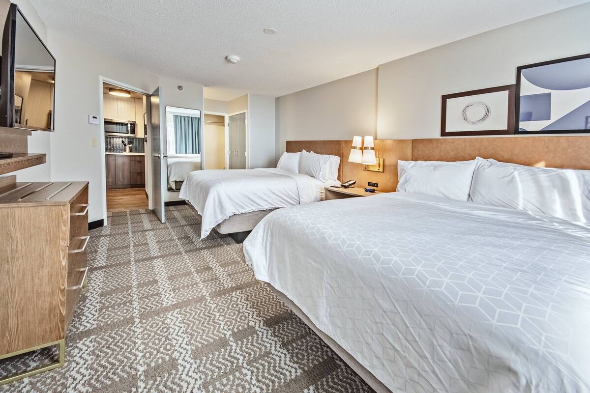 A hotel room with two beds, a desk, a mirror, a wall-mounted TV, a kitchen area, and a patterned rug.