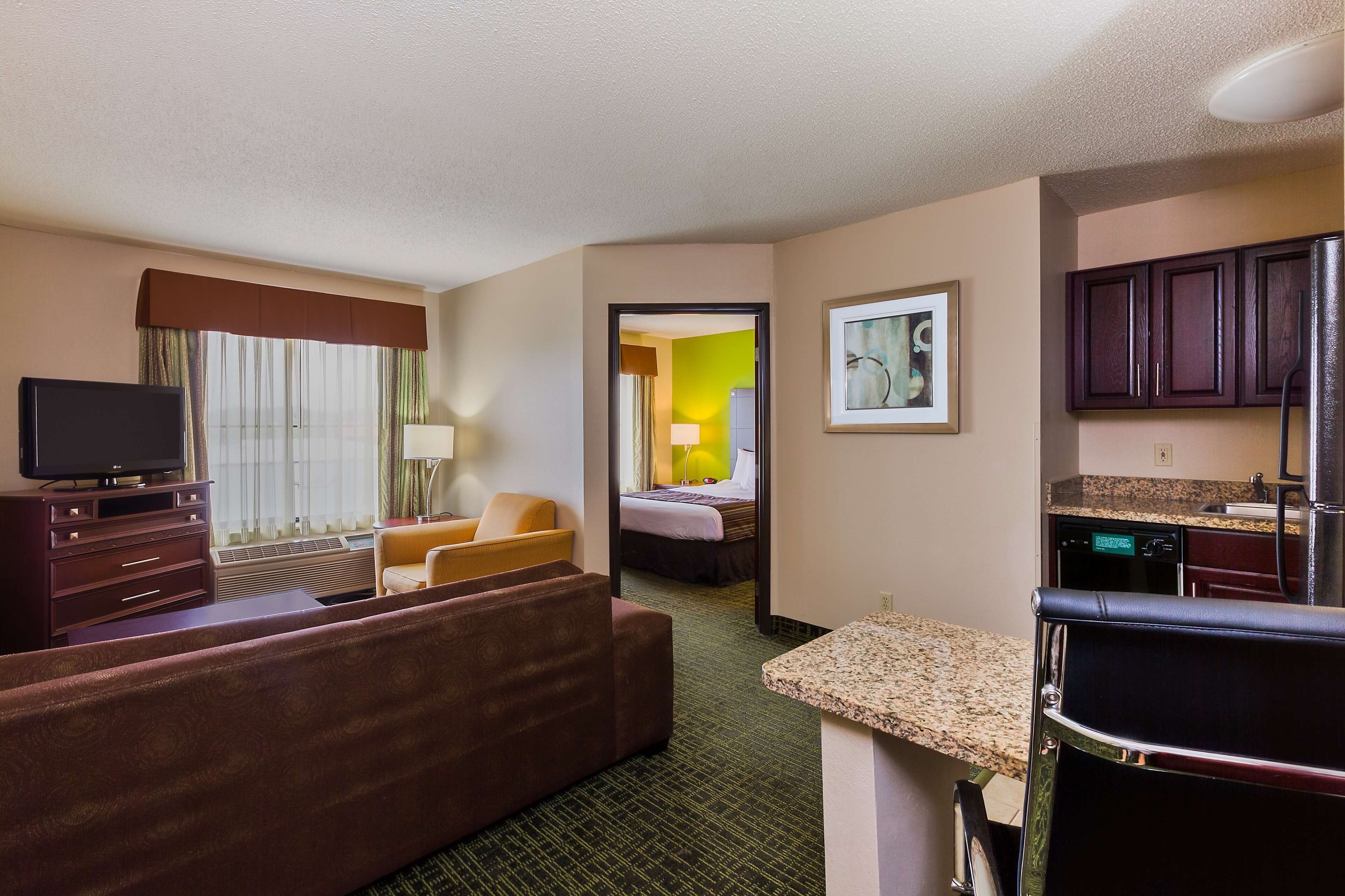 Quarto de hotel com televisão, sofá, poltrona, cama e cozinha compacta.