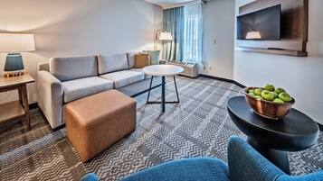 A modern hotel room with a grey sofa, a round coffee table, a brown ottoman, a blue armchair, and a bowl of green apples on a black side table.
