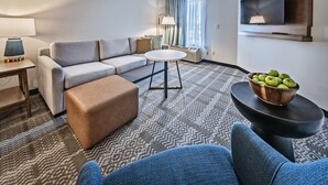 A modern hotel room with a grey sofa, a round coffee table, a small ottoman, a TV mounted on the wall, and a window with curtains. - Staybridge Suites Cedar Rapids North by IHG (Cedar Rapids)