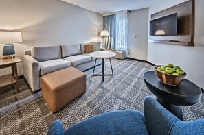 A modern hotel room with a grey sofa, a round coffee table, a small ottoman, a TV mounted on the wall, and a window with curtains. - Staybridge Suites Cedar Rapids North by IHG (Cedar Rapids)