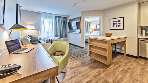 A hotel room with a wooden desk, a laptop, a green chair, a television, a dining area, and a kitchen area. - Staybridge Suites Cedar Rapids North by IHG (Cedar Rapids)
