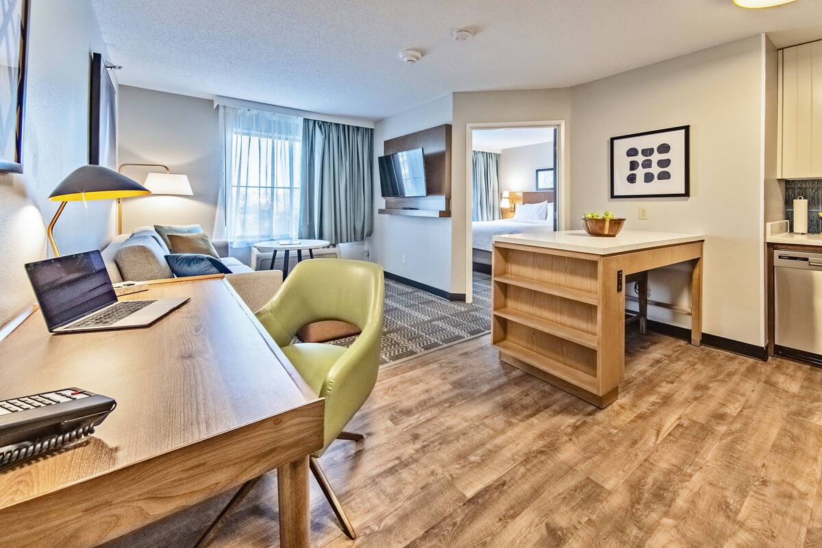 A hotel room with a wooden desk, a laptop, a green chair, a television, a dining area, and a kitchen area.