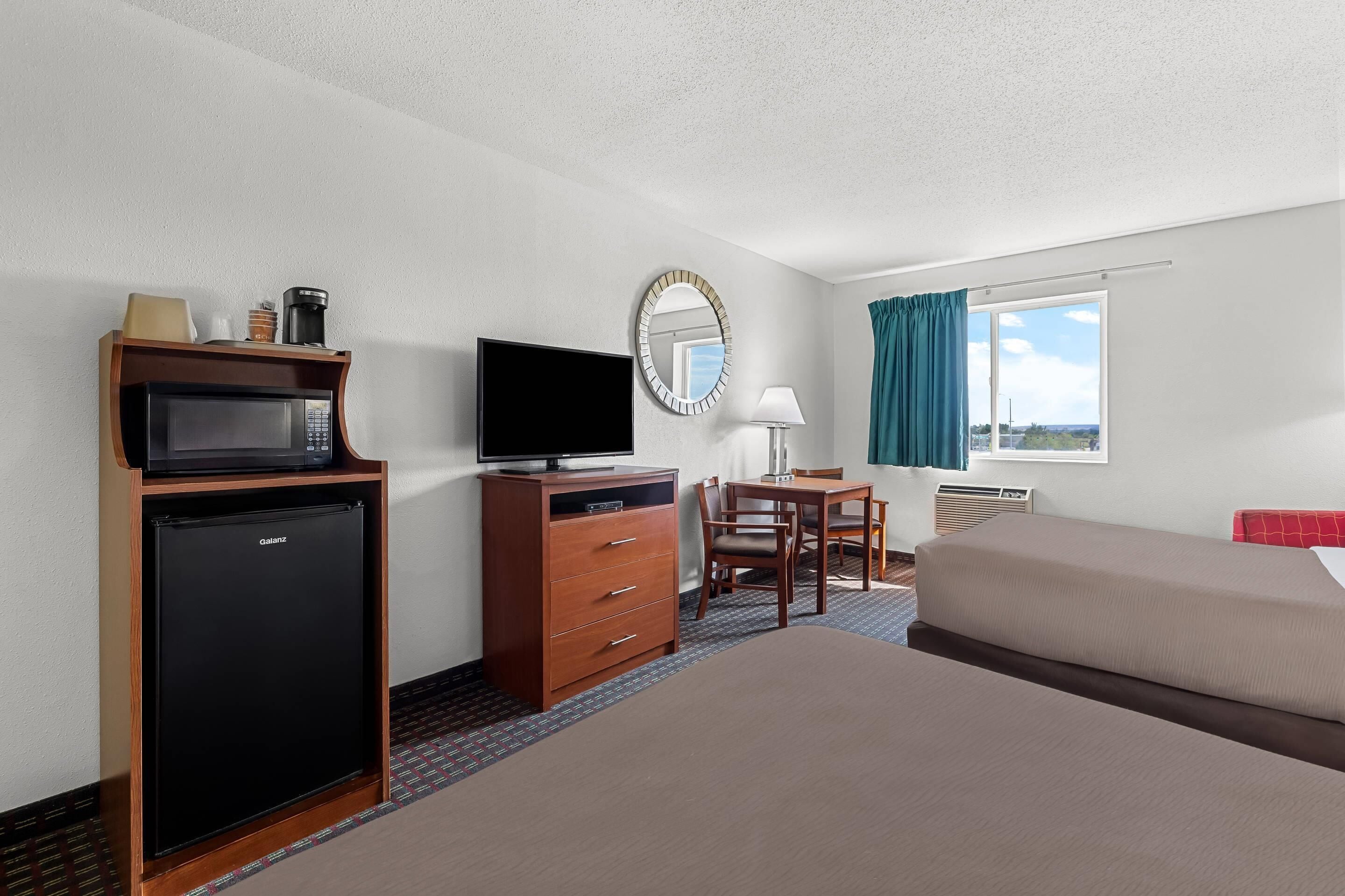 A hotel room with a bed, a desk with a chair, a television, a minibar, a mirror, and a window with curtains.